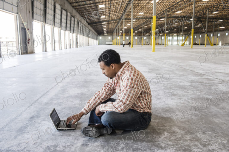 Black man working on lap top computer in front of loading dock doors in a new waerhouse.
