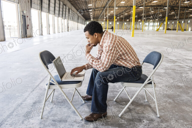 Black man working on lap top computer in front of loading dock doors in a new warehouse.