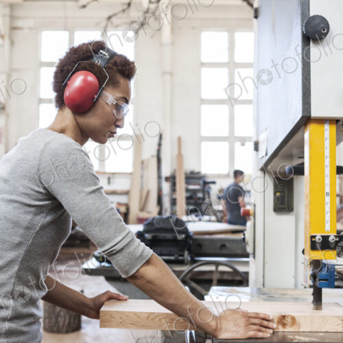 Black woman factory worker using a band saw to cut wood in a woodworking factory.