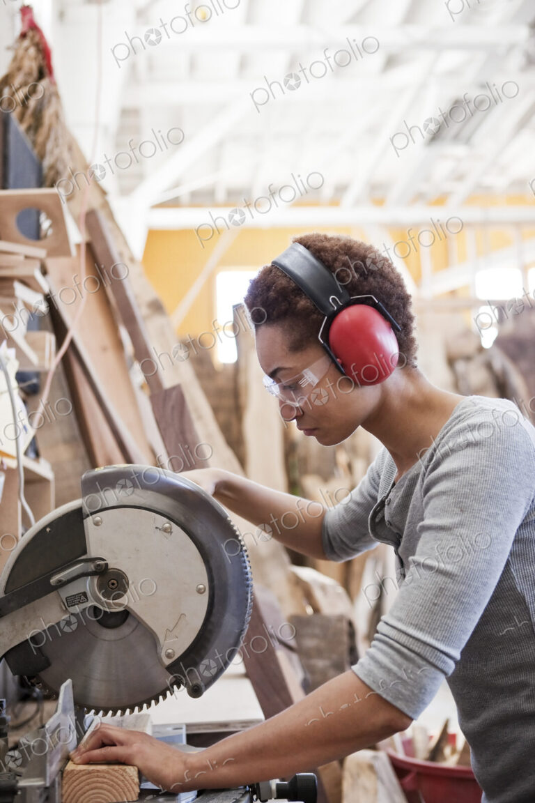 Black woman factory worker using a radial saw to cut wood in a woodworking factory.