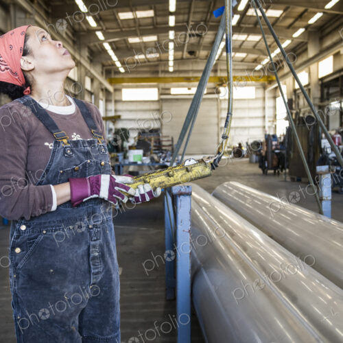 Black woman factory worker using an overhead lift to move pipes in a sheet metal factory.