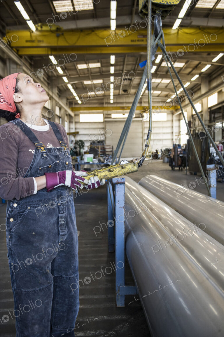 Black woman factory worker using an overhead lift to move pipes in a sheet metal factory.