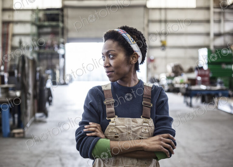 Black woman factory worker wearing coveralls in a large sheet metal factory.