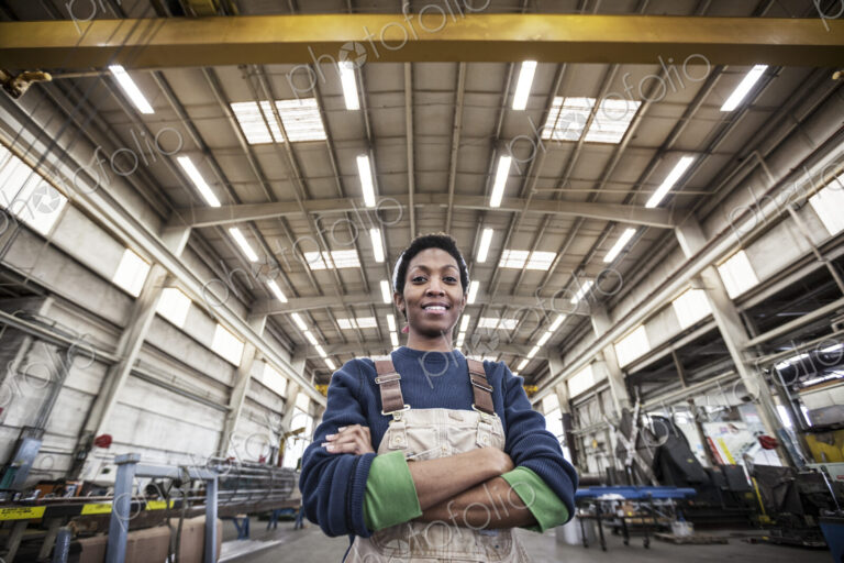Black woman factory worker wearing coveralls in a large sheet metal factory.