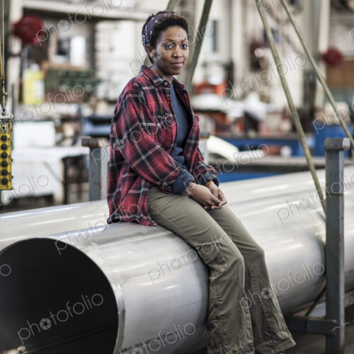 Black woman sitting next to a lift with aluminum pipes in a sheet metal factory.