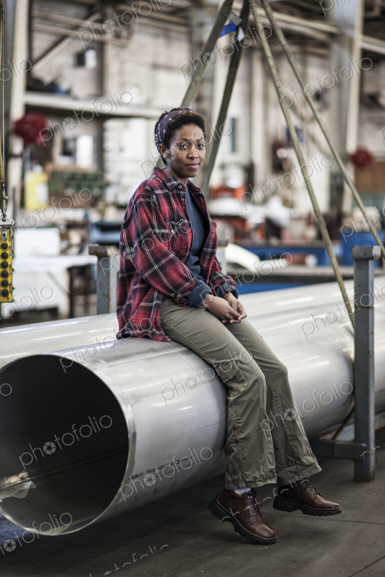 Black woman sitting next to a lift with aluminum pipes in a sheet metal factory.