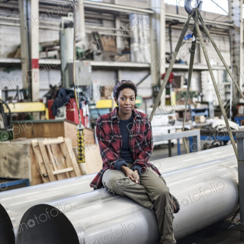 Black woman sitting next to a lift with aluminum pipes in a sheet metal factory.