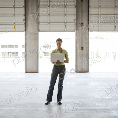 Black woman standing in front of new warehouse loading dock doors and using a lap top…