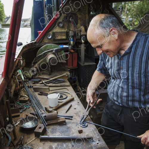 Blacksmith in his floating workshop on a narrowboat, holding pliers and metal rod,…