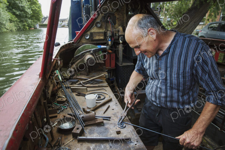 Blacksmith in his floating workshop on a narrowboat, holding pliers and metal rod,…