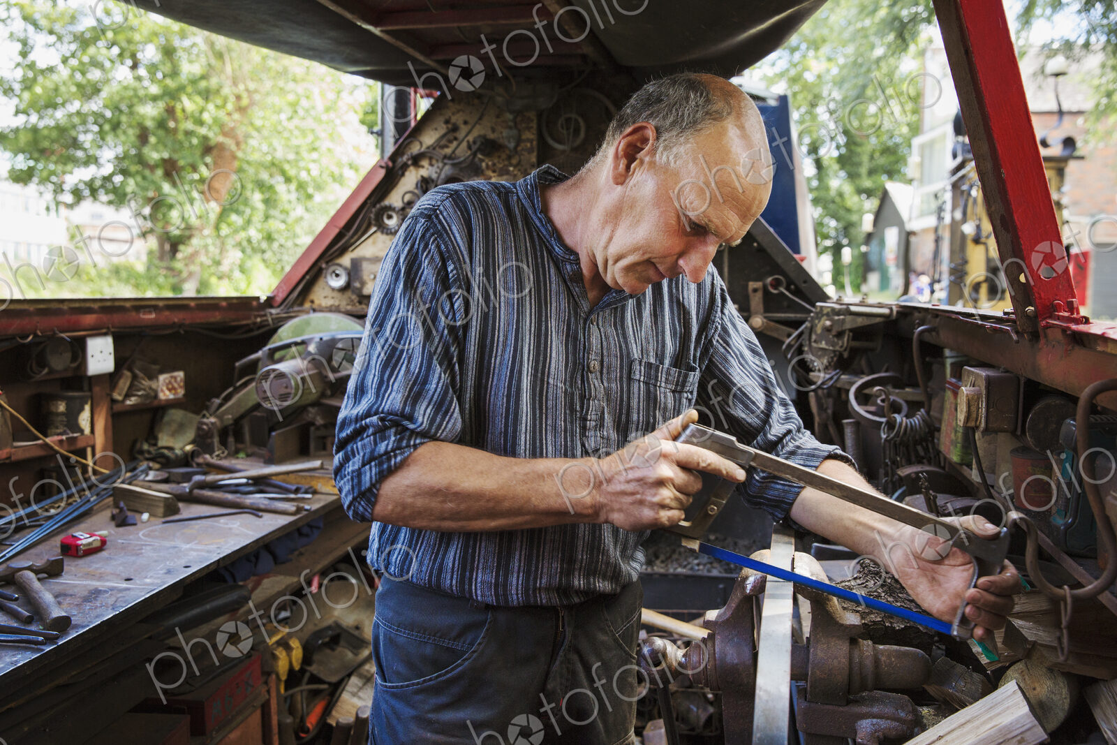 Blacksmith in his workshop on a working barge, at a work bench using a hacksaw.
