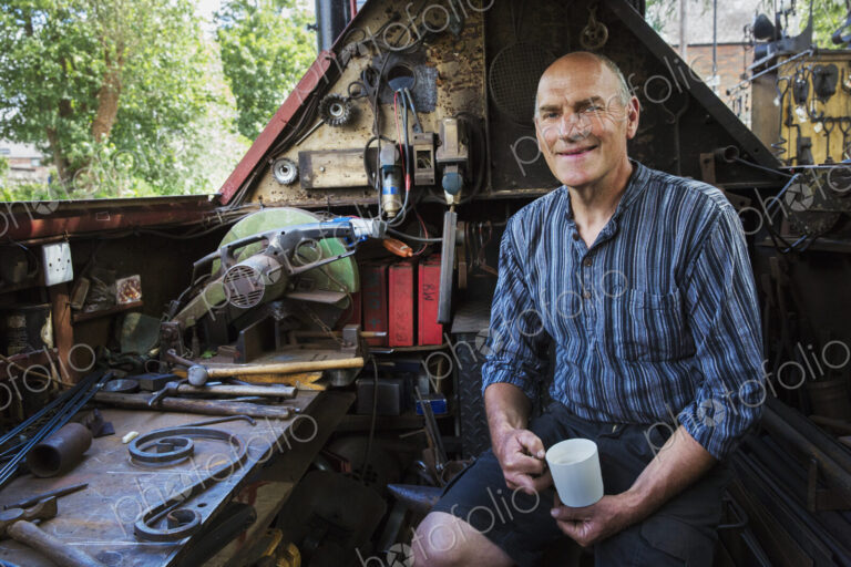 Blacksmith sitting on a working boat, a narrowboat on a waterway, holding a mug,…