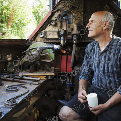 Blacksmith sitting on a working narrowboat on a waterway, in his workshop holding mug. Blacksmith sitting on a working narrowboat on a waterway, in his workshop holding mug.