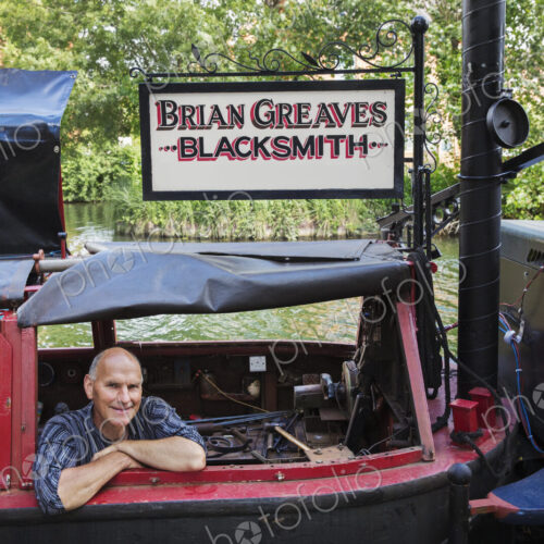 Blacksmith sitting on his working narrowboat on a waterway, looking at camera, smiling. Blacksmith sitting on his working narrowboat on a waterway, looking at camera, smiling.