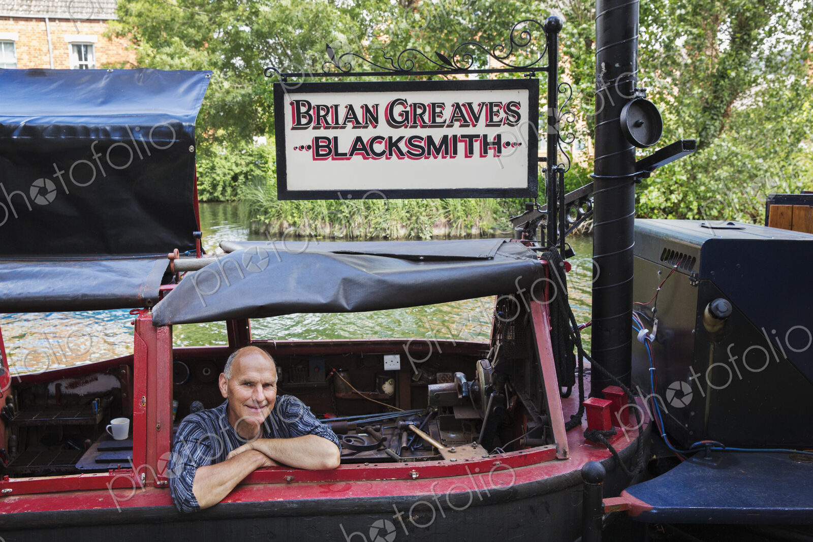 Blacksmith sitting on his working narrowboat on a waterway, looking at camera, smiling.