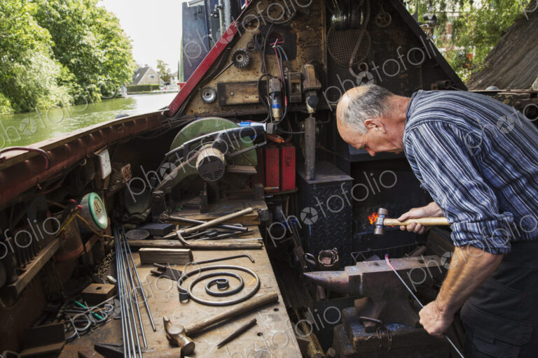 Blacksmith working in a small space on his narrowboat, a barge on river, bending over…