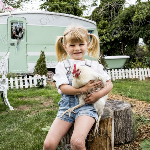 Blond girl sitting in garden holding white chicken, white and green retro caravan in… Blond girl sitting in garden holding white chicken, white and green retro caravan in…