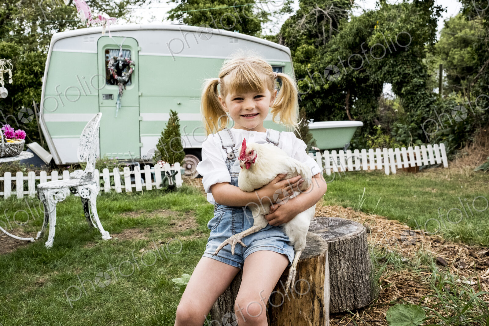 Blond girl sitting in garden holding white chicken, white and green retro caravan in…
