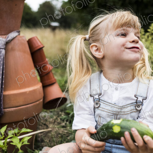 Blond girl sitting in garden next to terracotta scarecrow, eating cucumber. Blond girl sitting in garden next to terracotta scarecrow, eating cucumber.