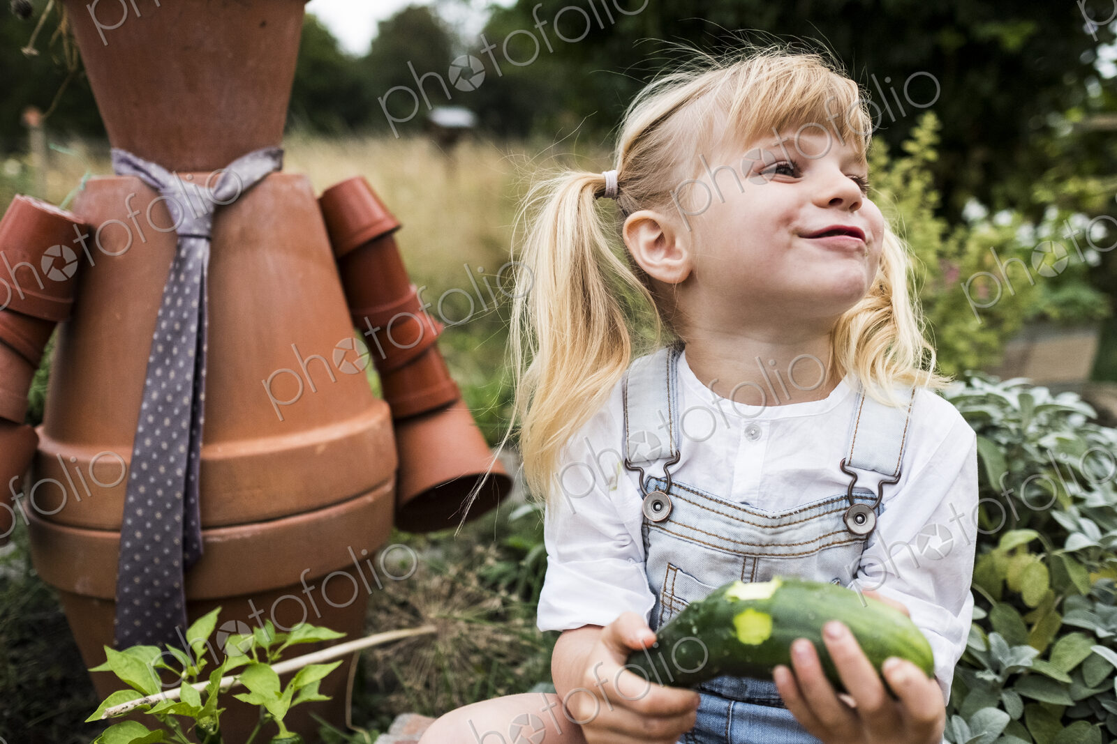 Blond girl sitting in garden next to terracotta scarecrow, eating cucumber.