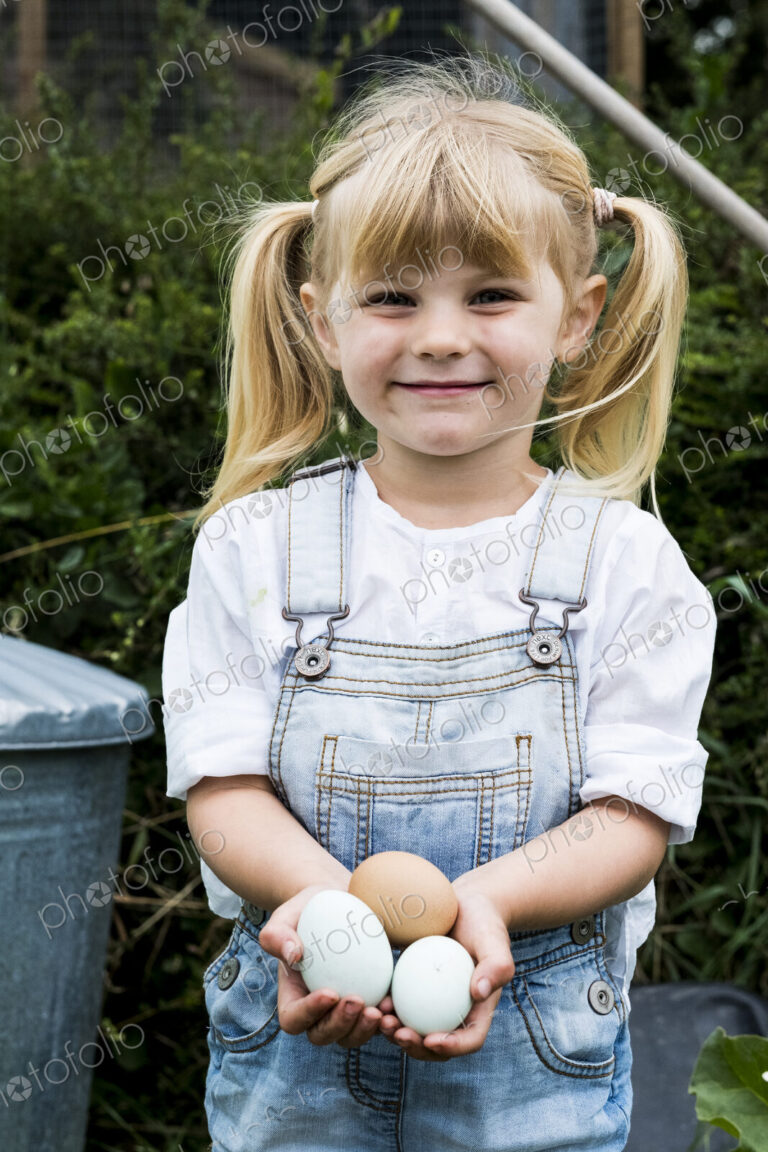 Blond girl standing in a garden, holding fresh eggs, smiling at camera.