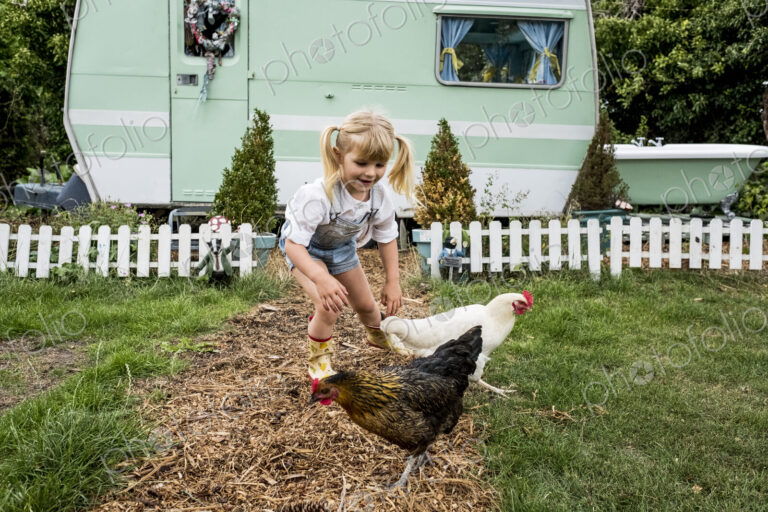 Blond girl with two chickens on a garden path, white and green retro caravan in…
