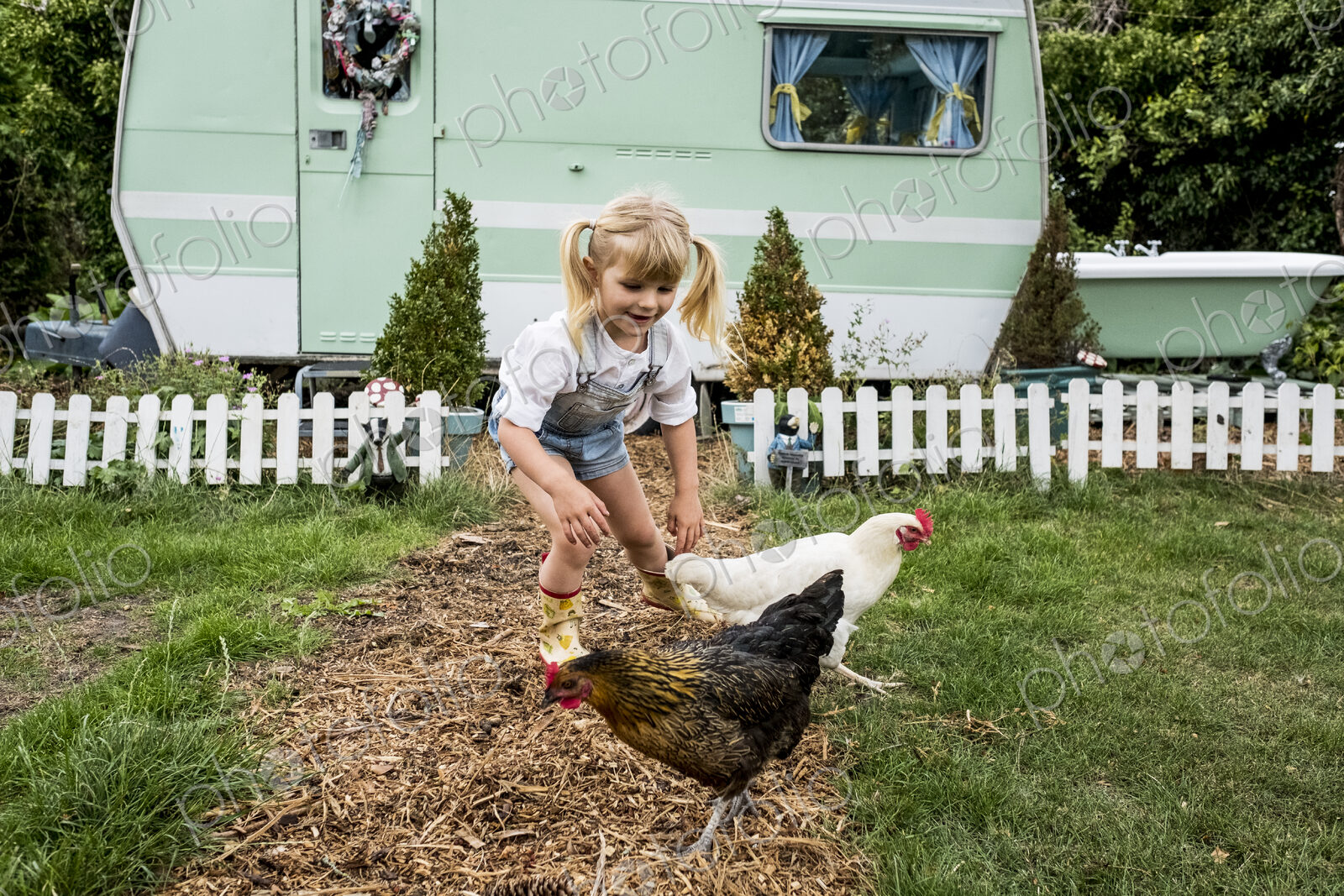 Blond girl with two chickens on a garden path, white and green retro caravan in…