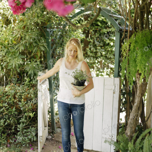 Blond woman entering a garden through a white wooden gate, carrying a plant pot. Blond woman entering a garden through a white wooden gate, carrying a plant pot.