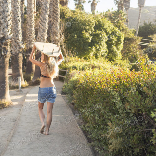 Blond woman in a black bikini and denim shorts carrying a surfboard on a path lined… Blond woman in a black bikini and denim shorts carrying a surfboard on a path lined…