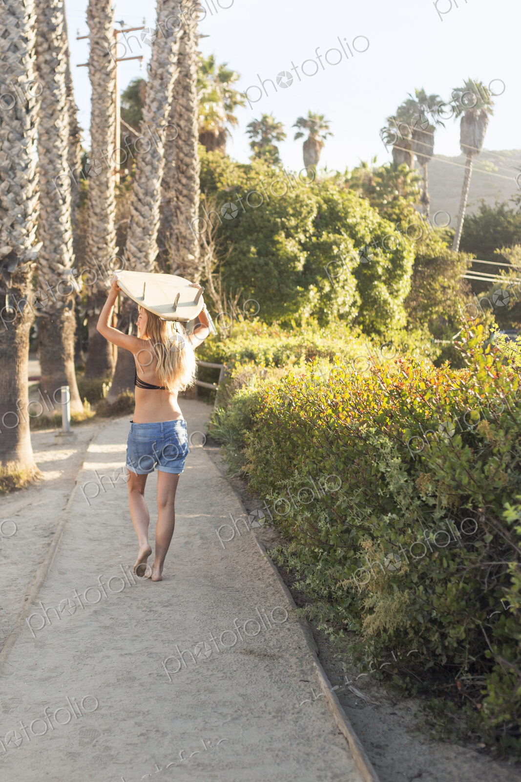 Blond woman in a black bikini and denim shorts carrying a surfboard on a path lined…