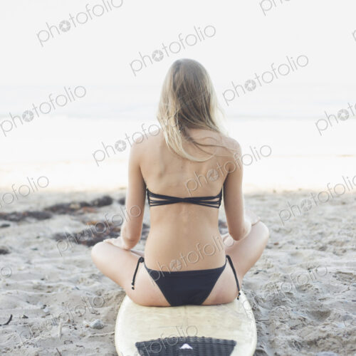 Blond woman in a black bikini sitting on a surfboard on a sandy beach. Blond woman in a black bikini sitting on a surfboard on a sandy beach.