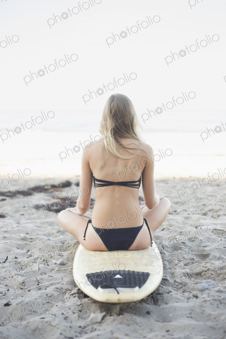 Blond woman in a black bikini sitting on a surfboard on a sandy beach.