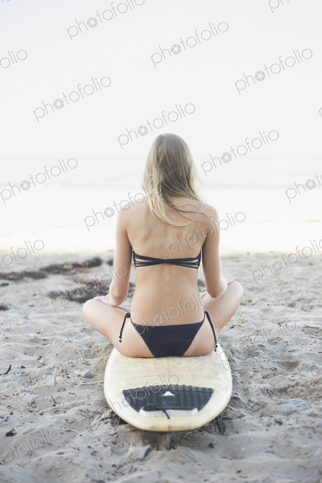 Blond woman in a black bikini sitting on a surfboard on a sandy beach.