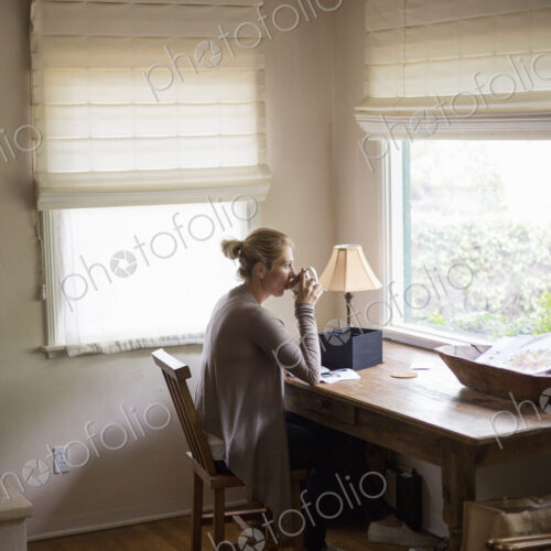 Blond woman sitting at a desk by a window, drinking a cup of tea. Blond woman sitting at a desk by a window, drinking a cup of tea.