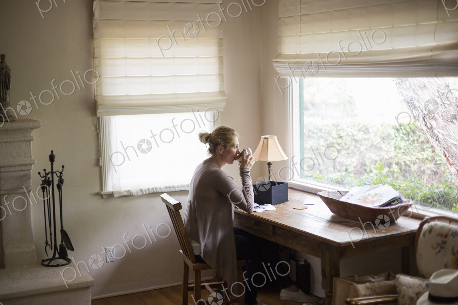 Blond woman sitting at a desk by a window, drinking a cup of tea.