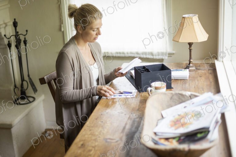 Blond woman sitting at a desk by a window, looking at photographs.