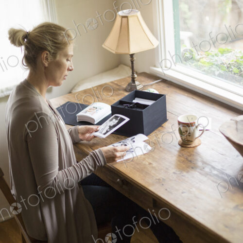 Blond woman sitting at a desk by a window, looking at photographs.