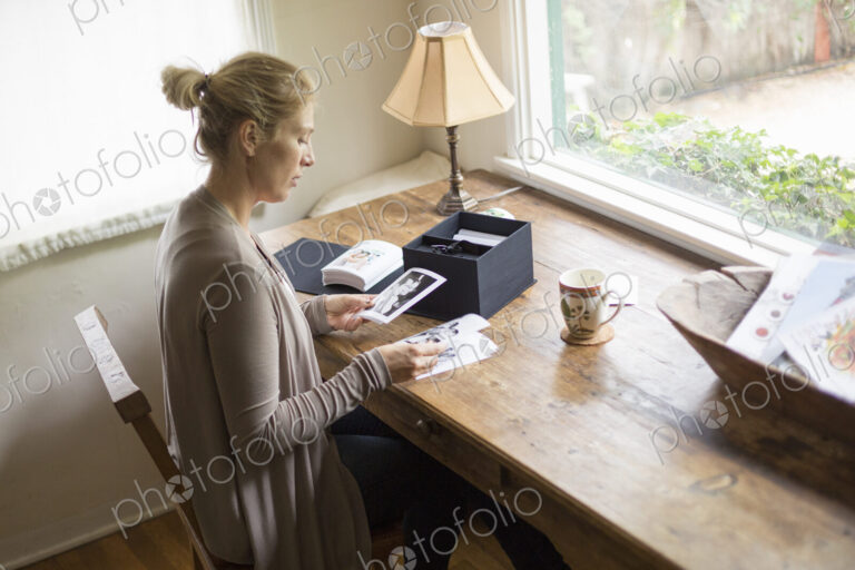 Blond woman sitting at a desk by a window, looking at photographs.