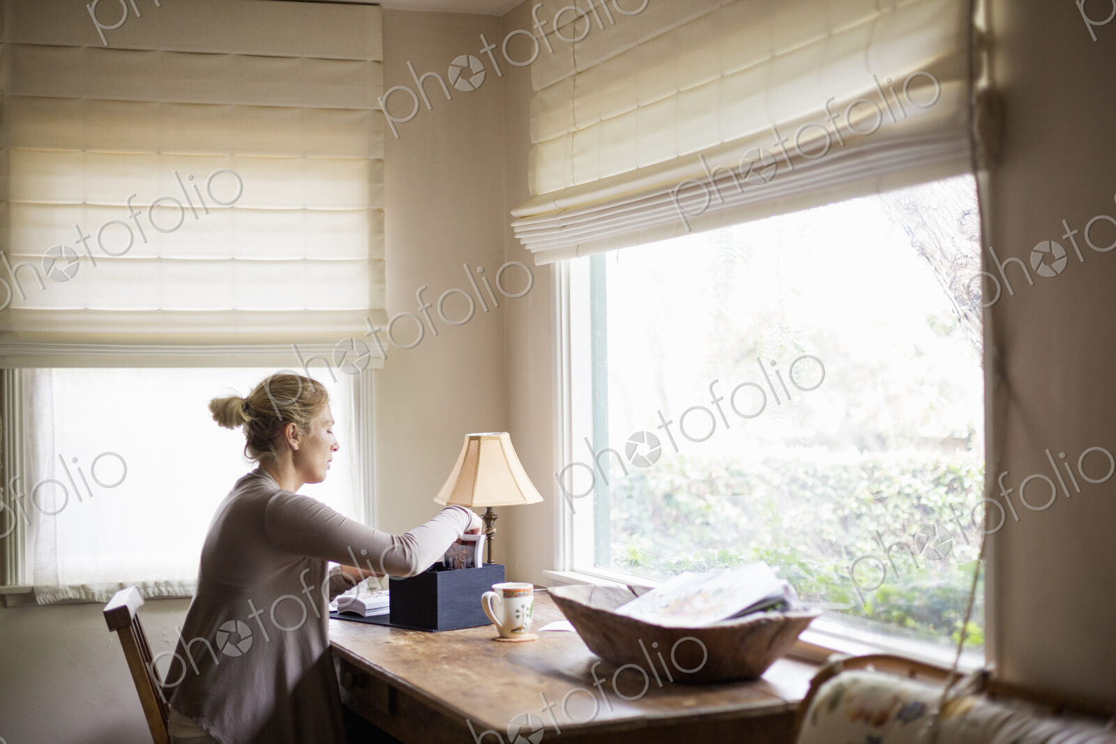 Blond woman sitting at a desk by a window, sorting through a box with photographs.