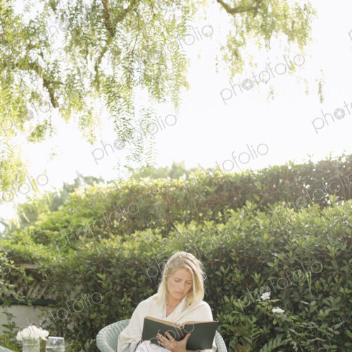 Blond woman sitting in a wicker chair in a garden, reading.