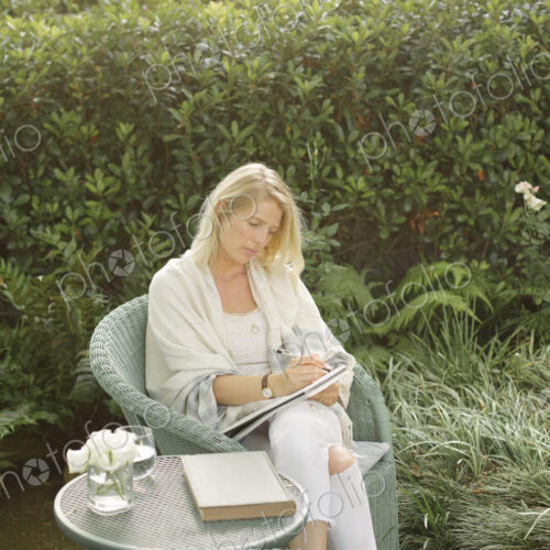 Blond woman sitting in a wicker chair in a garden, writing.