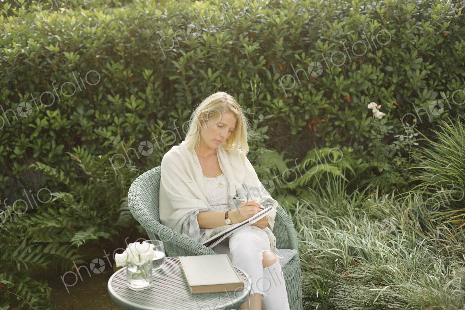 Blond woman sitting in a wicker chair in a garden, writing.