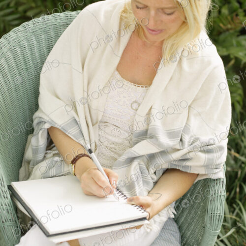 Blond woman sitting in a wicker chair in a garden, writing.