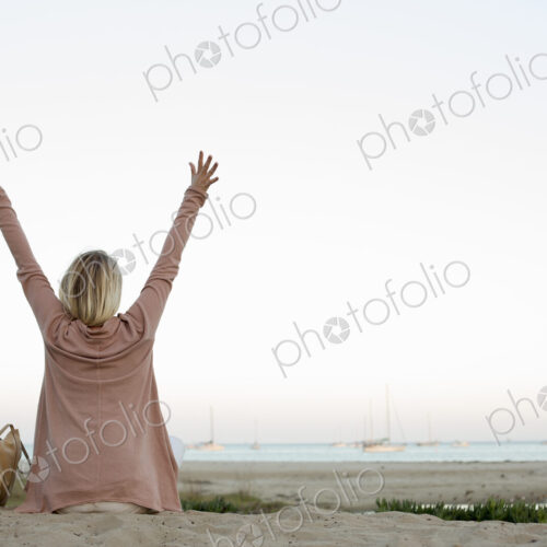 Blond woman sitting on a sandy beach, arms raised. Blond woman sitting on a sandy beach, arms raised.