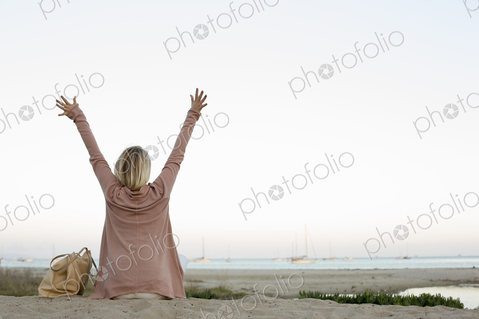 Blond woman sitting on a sandy beach, arms raised.