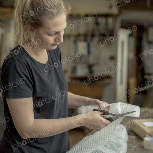 Blond woman standing in a workshop, cutting bubble wrap with pair of scissors,… Blond woman standing in a workshop, cutting bubble wrap with pair of scissors,…