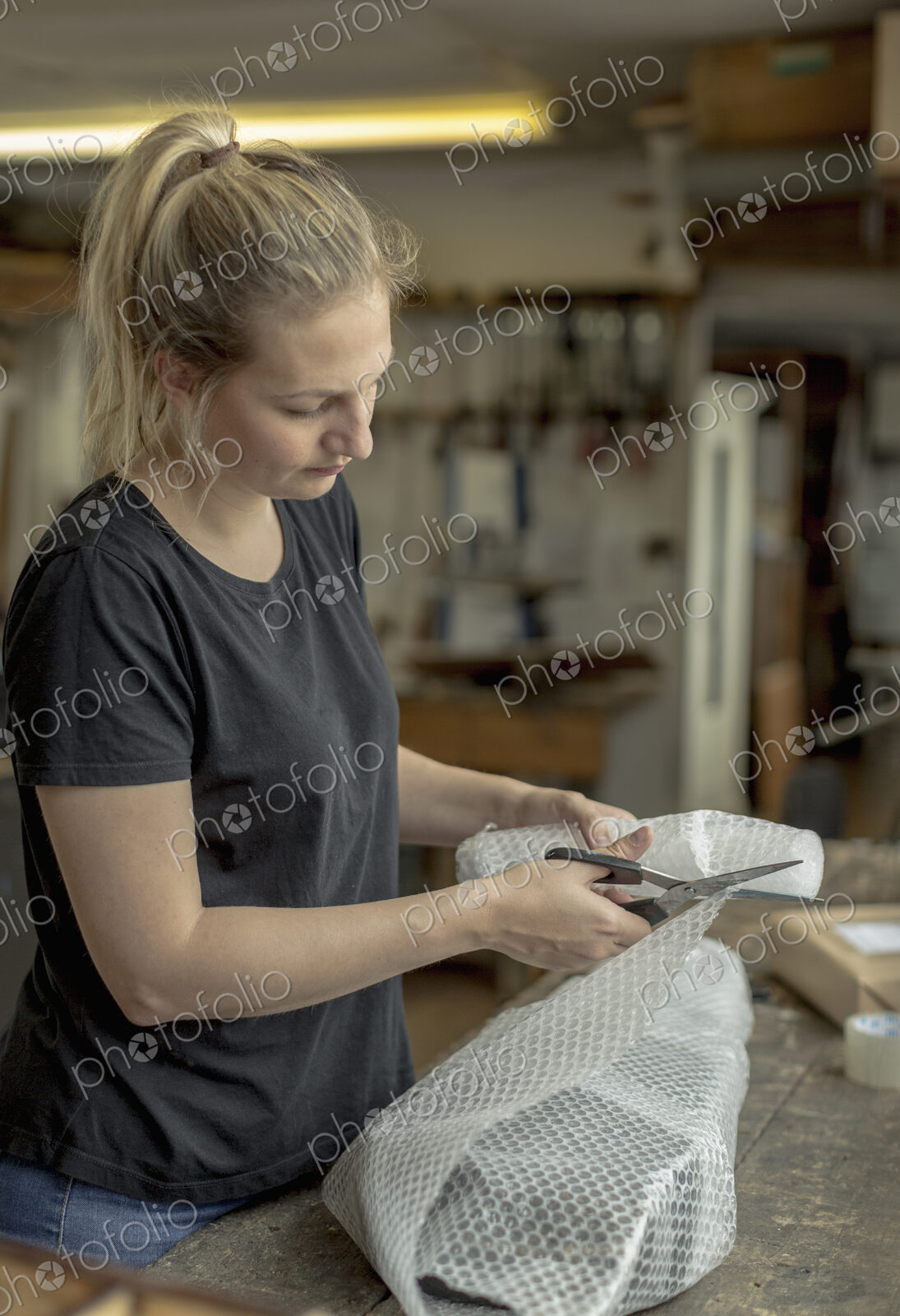 Blond woman standing in a workshop, cutting bubble wrap with pair of scissors,…