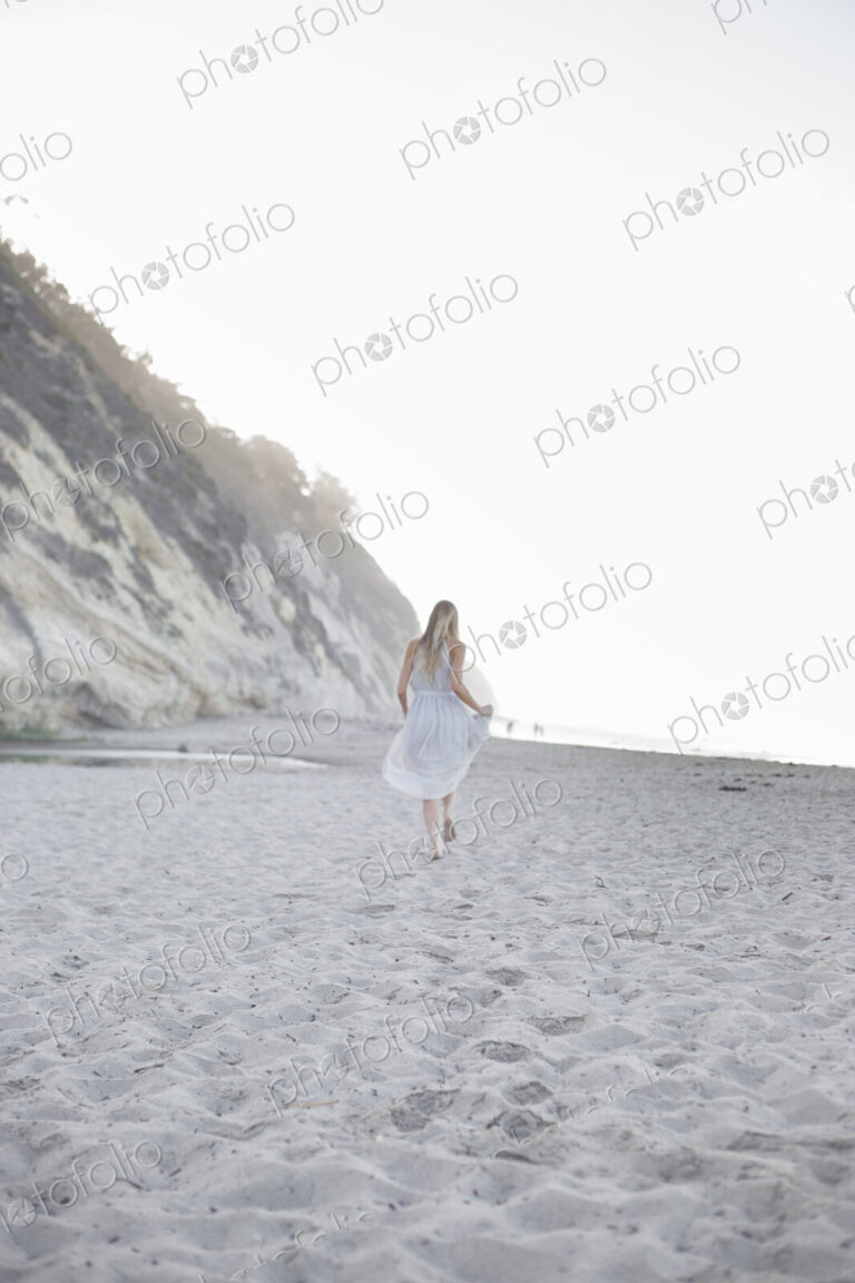 Blond woman walking on a sandy beach near a cliff.