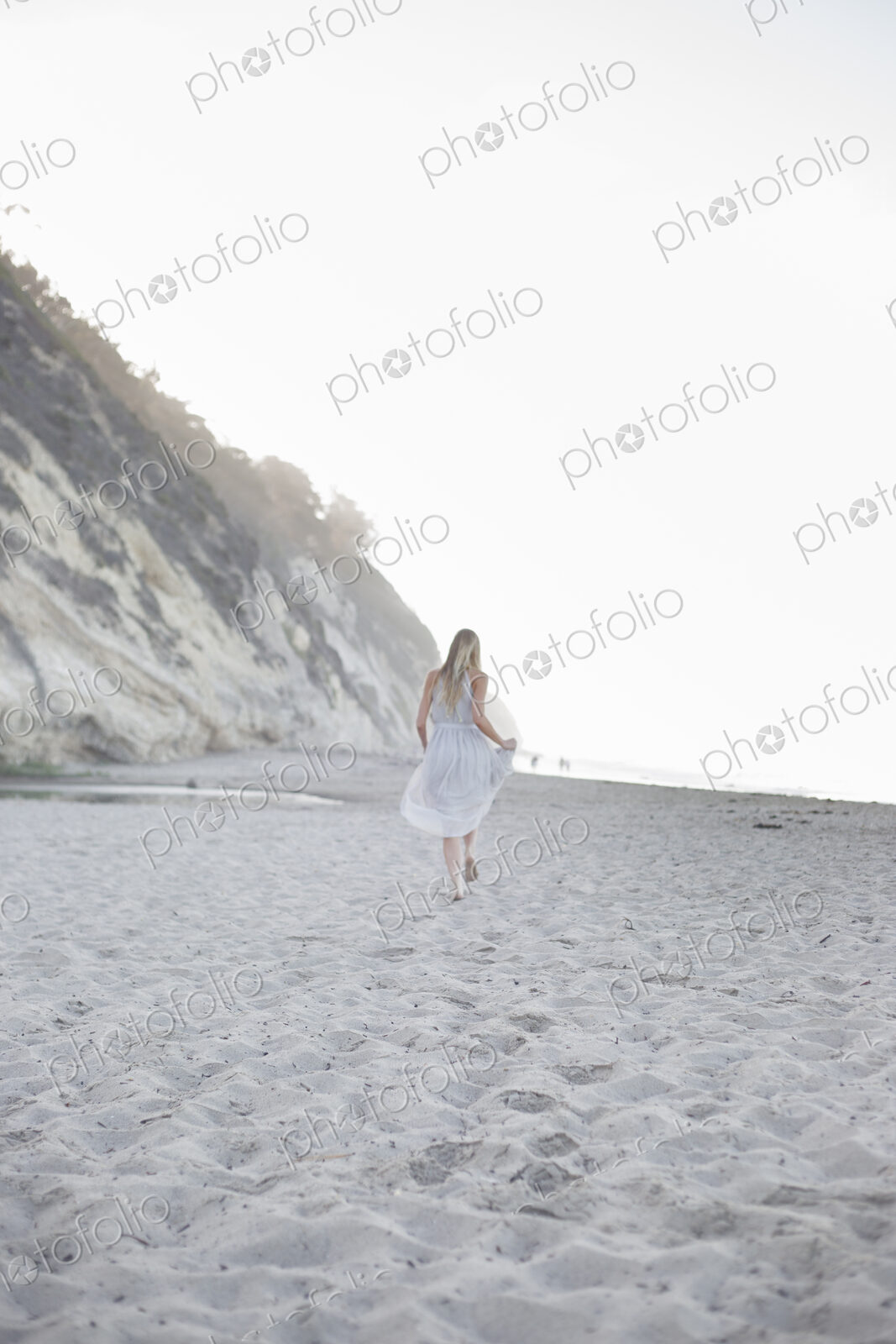 Blond woman walking on a sandy beach near a cliff.