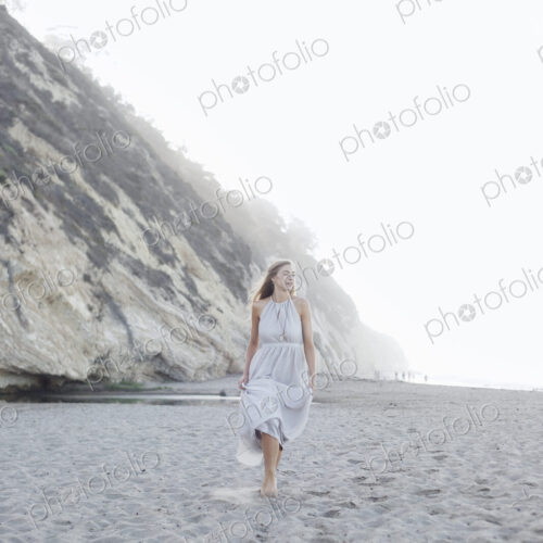Blond woman walking on a sandy beach near a cliff.